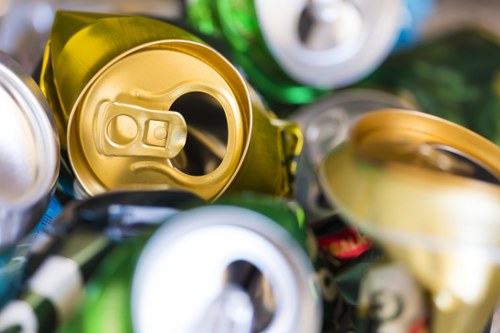 Close-up image of various crushed and partially opened aluminium beverage cans in different metallic colours including gold, silver, green, and blue, piled together on a surface, possibly for recycling or waste disposal. The cans have textured, ridged surfaces, with some showing familiar pull-tabs and indentations from opening. The background is slightly blurred, emphasizing the cans in the foreground, and the lighting highlights the reflective surfaces of the aluminium materials. This collection of cans is representative of typical household or outdoor waste that a rubbish removal service such as Garden Clearance Edmonton might handle, particularly in the context of recycling or waste collection for properties around Edmonton or nearby locations in North London, aligning with outdoor waste clearance activities in the local area.