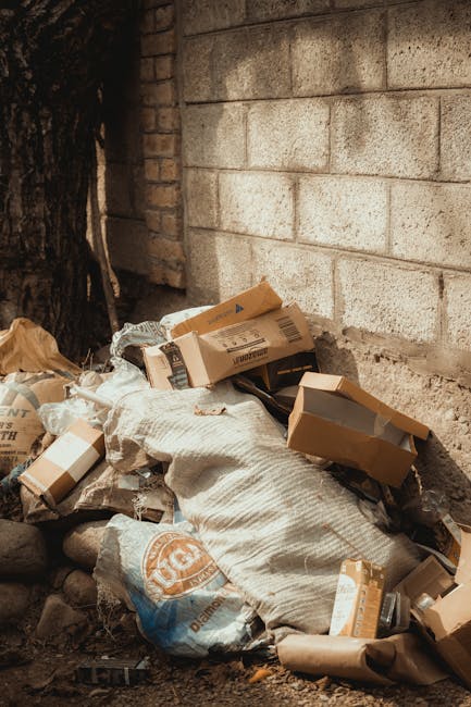 A white urban rubbish collection truck with a large, open rear compactor area is positioned on a cobbled street next to a row of older, multi-story buildings with a weathered facade. The truck's surfa