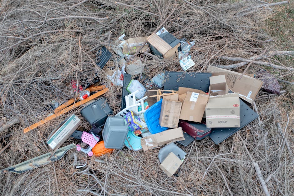 A green, weathered clothing donation bin positioned outdoors on a pavement, with a clutter of discarded items including shoes, clothing, bags, and various small objects scattered around its base. The 