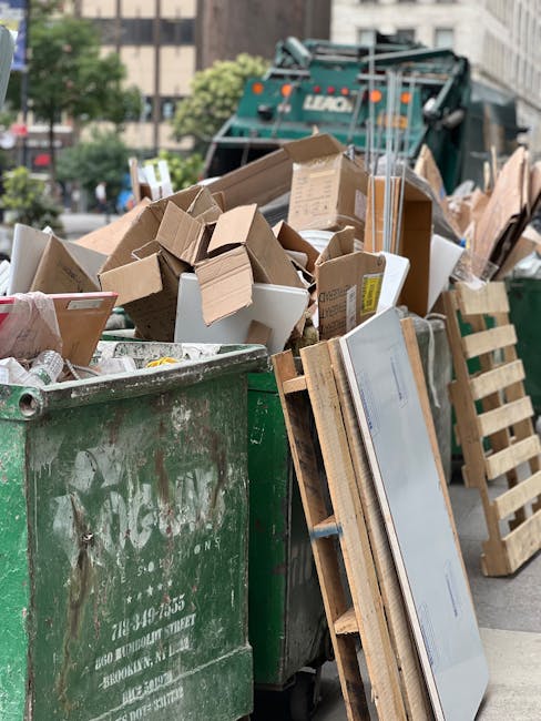 A white waste collection truck is parked on a cobblestone street beside a row of multi-storey buildings with aged facades, some showing peeling paint. The truck's rear hopper is open, revealing rusted